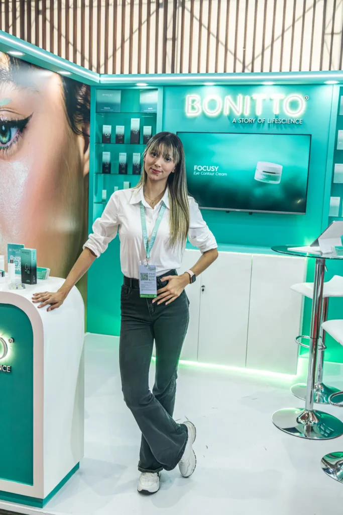 امرأة ترتدي قميصاً أبيض وسروالاً رمادياً وتقف في كشك BONITTO للعناية بالبشرة باللونين الأبيض والأزرق المخضر، وتبتسم للكاميرا، مع عرض المنتج وصورة كبيرة للعين في الخلفية.