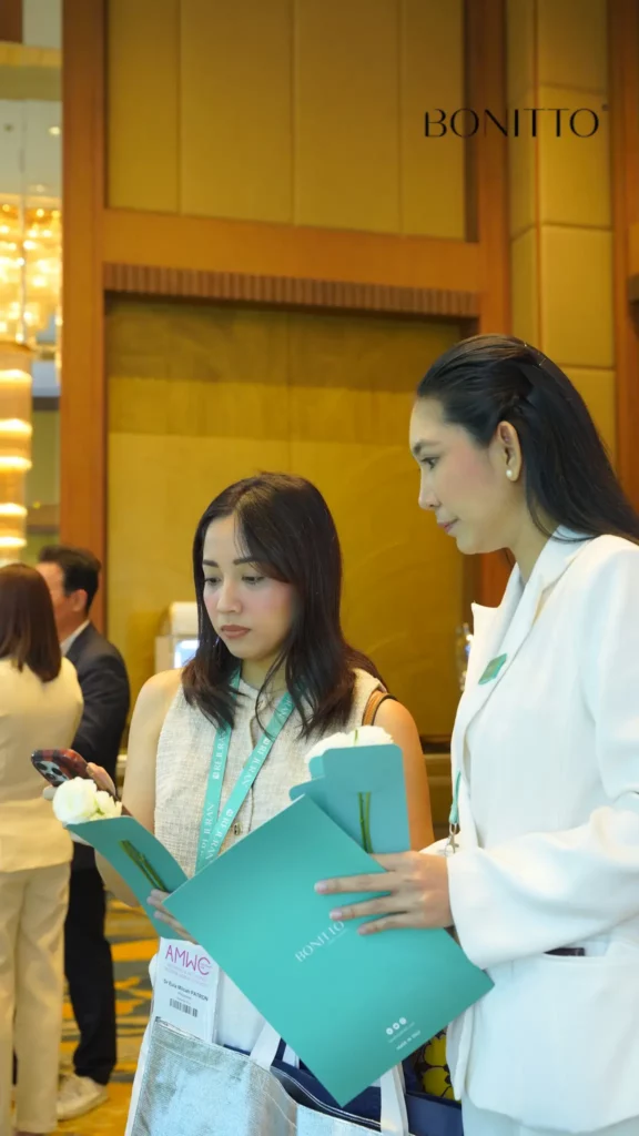 Two women standing indoors at an event, holding teal folders and wearing lanyards. One woman looks at her phone while the other looks at her folder. A "BONITTO" logo is visible in the upper right corner.
