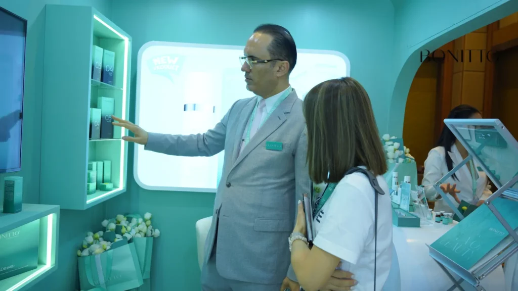 A man in a light gray suit shows skincare products on a shelf to a woman with brown hair at a turquoise-colored exhibition booth. A glowing display and product samples are visible in the background.