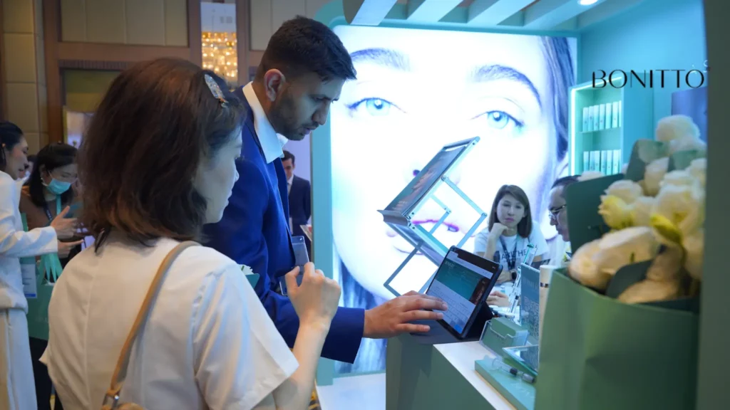 A man in a blue suit demonstrates a tablet to a woman at a modern booth with a large illuminated image of a face in the background and the word "BONITTO" on the wall. Other people stand and sit nearby.