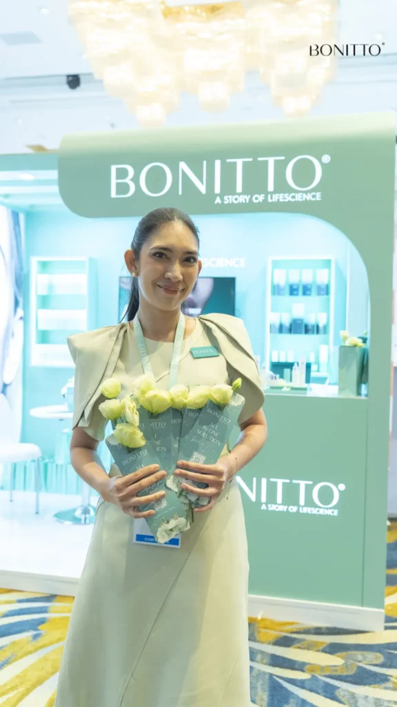 A woman in a beige dress stands indoors holding several bouquets of white roses. She is smiling in front of a turquoise BONITTO display booth with skincare products visible in the background.