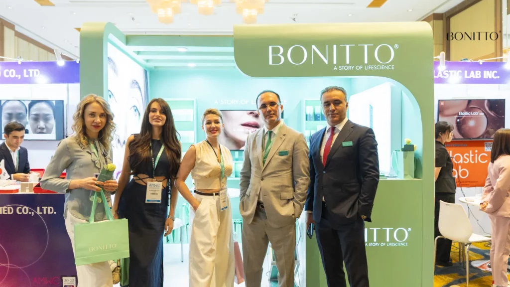 Five professionally dressed people stand and smile at a BONITTO booth at an event or expo. The booth features a mint green color and branding, with product displays and informational material in the background.