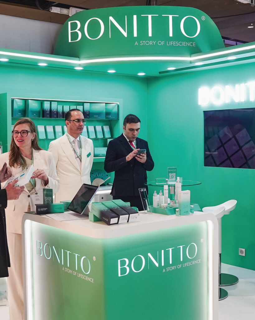 Three people stand at a bright green BONITTO booth at an event, with product displays and promotional materials on the counter and shelves behind them. Two people wear white suits and one wears a black suit.