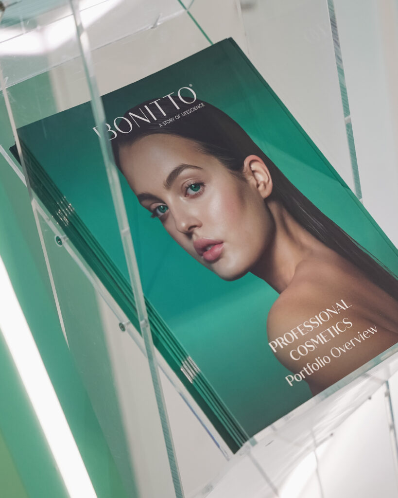 A stack of green Bonitto "Professional Cosmetics Portfolio Overview" brochures sits on a clear acrylic shelf, featuring a woman with flawless makeup on the cover.
