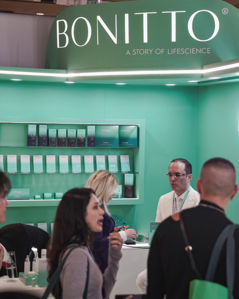 People gather at a turquoise BONITTO booth displaying skincare products, with shelves of boxes behind two staff members engaging with visitors under a large sign reading “A STORY OF LIFESCIENCE.”.
