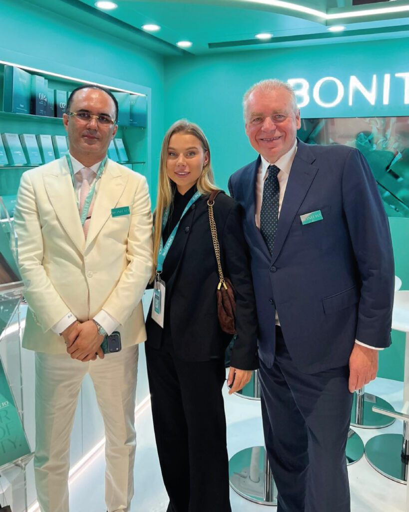 Three people in formal business attire stand together, smiling, inside a modern, teal-colored exhibition booth with product displays and the word "BONIT" partially visible on the wall behind them.
