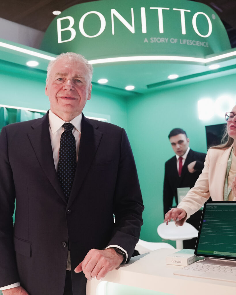 A man in a suit stands smiling at a booth with a green backdrop displaying "BONITTO A STORY OF LIFESCIENCE." Two people are behind a counter with a laptop and product packaging visible.