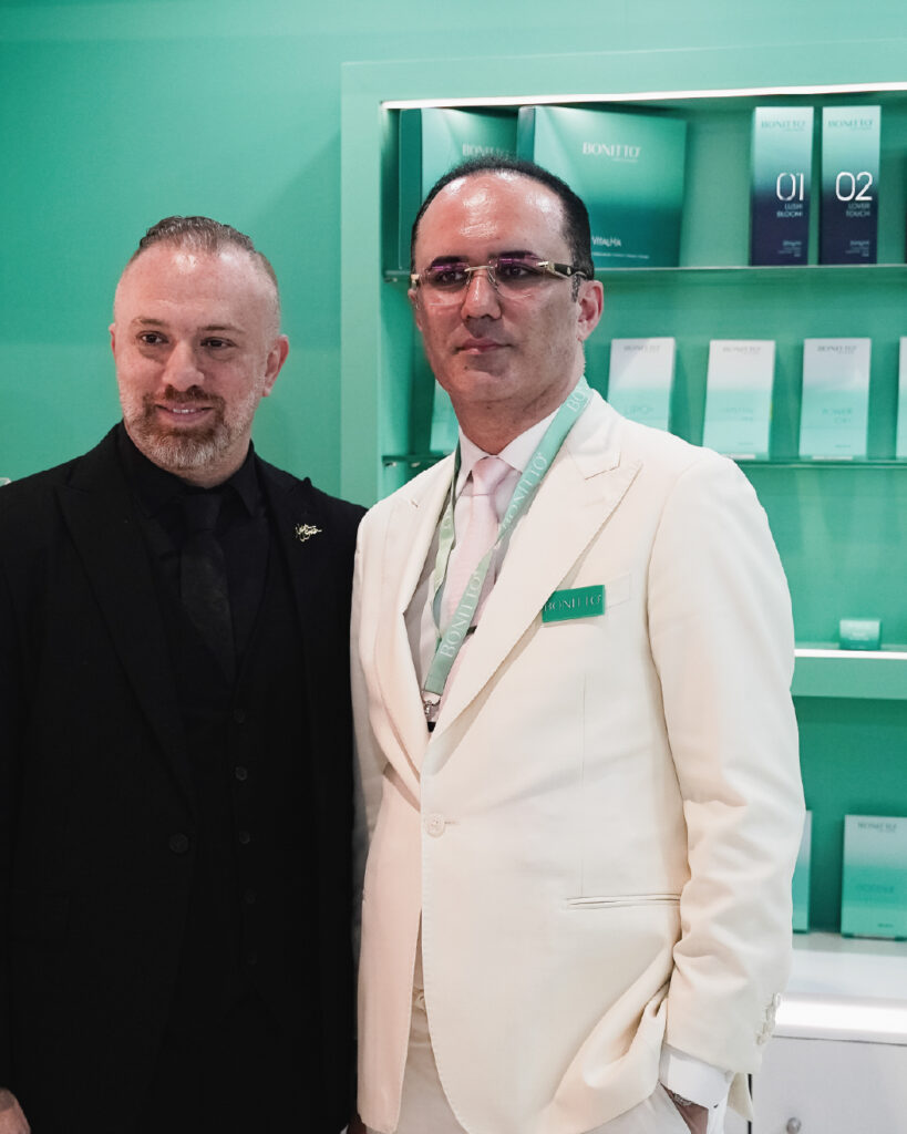 Two men in suits, one in black and one in white, stand together in front of green shelves displaying BONIQU skincare products. The background and product packaging are predominantly green.