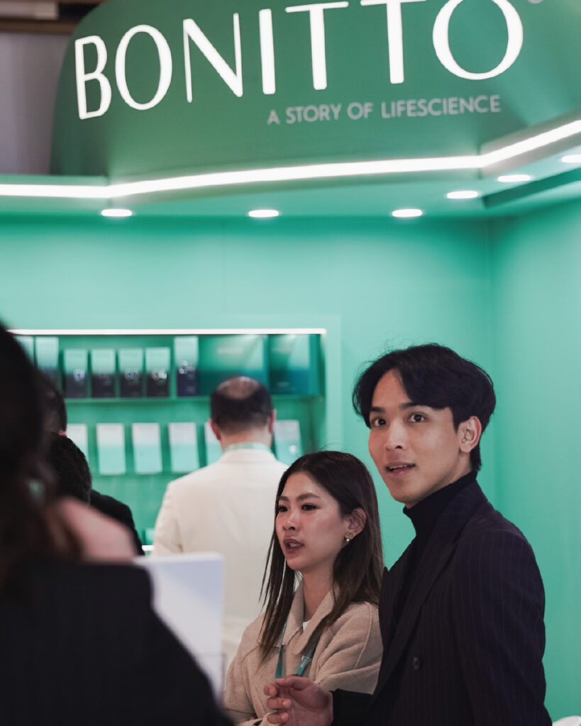 Two people stand in front of a green booth with the sign "BONITTO: A Story of Lifescience" above them. They appear to be interacting with others at the booth, which displays products on shelves behind them.