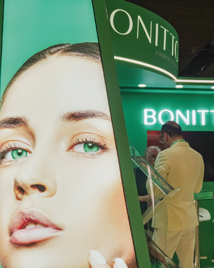 A large advertisement featuring a close-up of a woman with green eyes is displayed at a green and white BONITTO booth, where two men in light suits are engaged in conversation in the background.