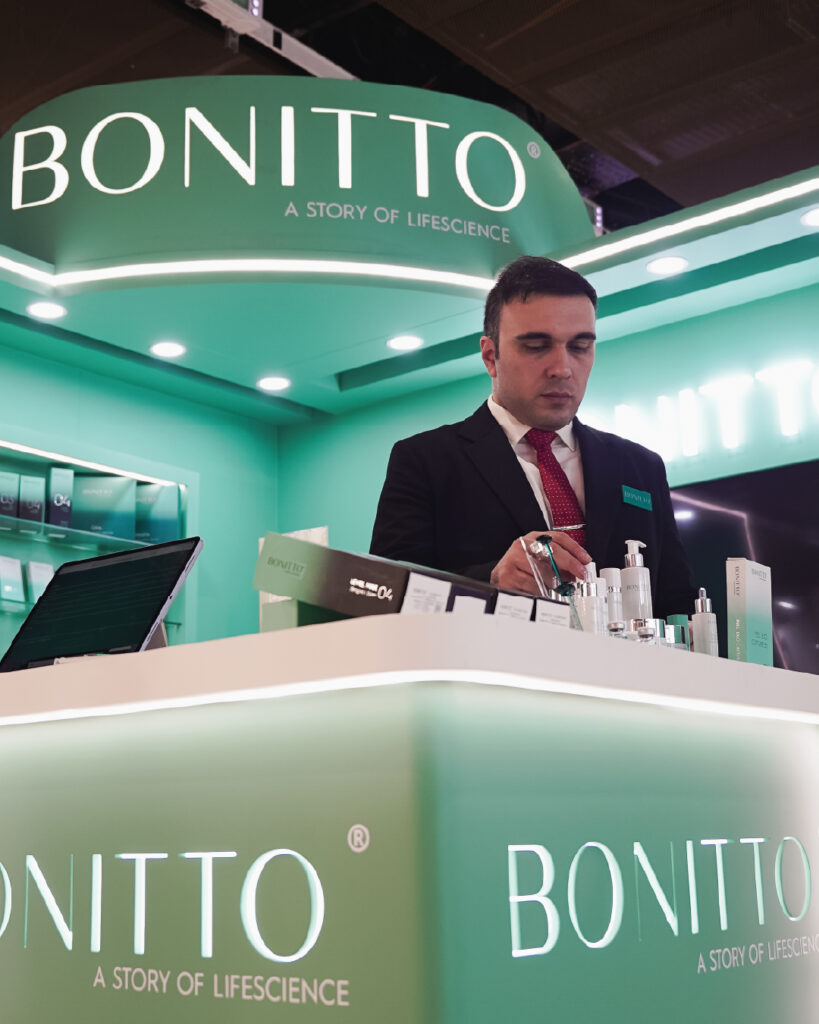 A man in a suit stands at a green and white BONITTO skincare display booth, arranging products under bright lights, with shelves of cosmetics behind him.